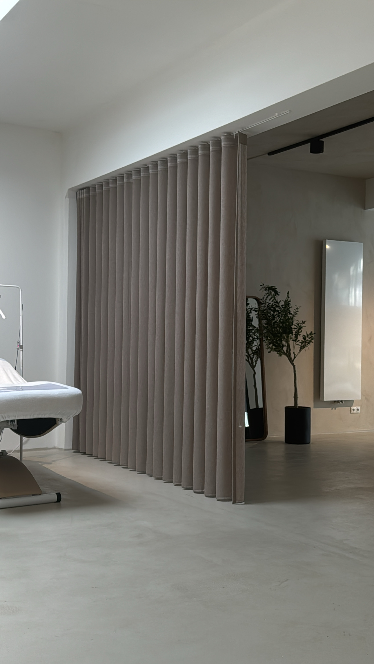 Behandelruimte met een behandelbed achter een taupe gordijn en een plant